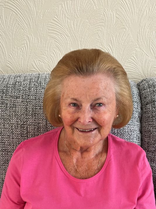 A picture of Carol, an older lady supporter of the charity, smiling at the camera, wearing a pink top, sat against a grey sofa.