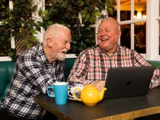 Two men smiling and laughing as they drink coffee in front of a laptop
